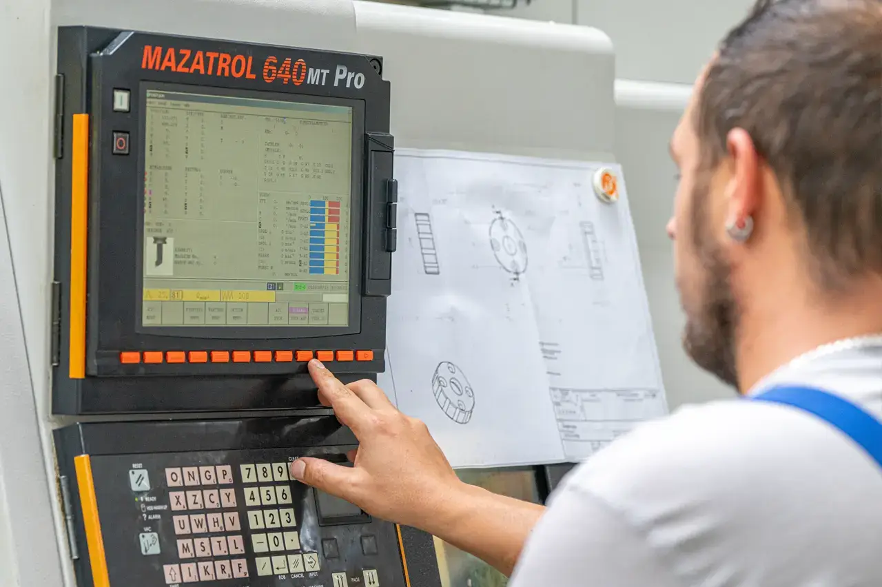 Mitarbeiter bedient die Steuerung einer CNC-Maschine, daneben hängt die Werkstückzeichnung
